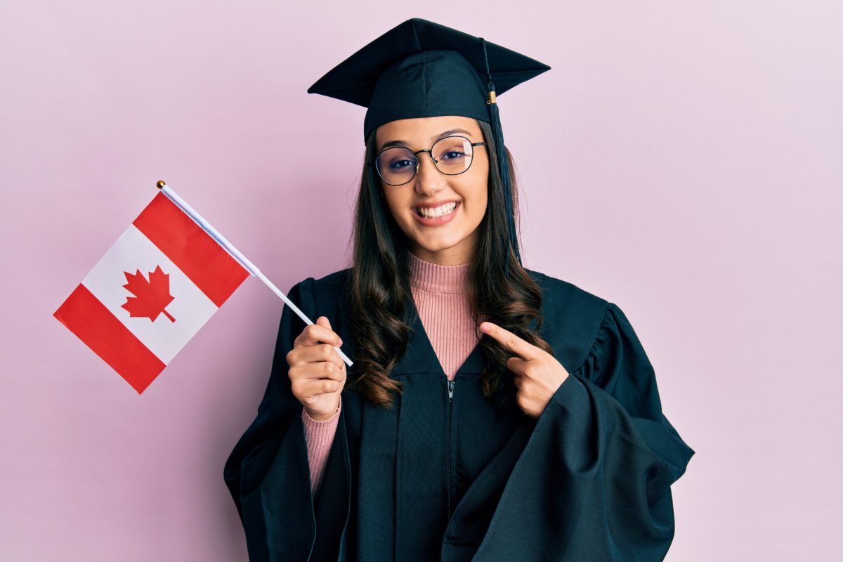 Young hispanic woman wearing graduation uniform holding canada flag